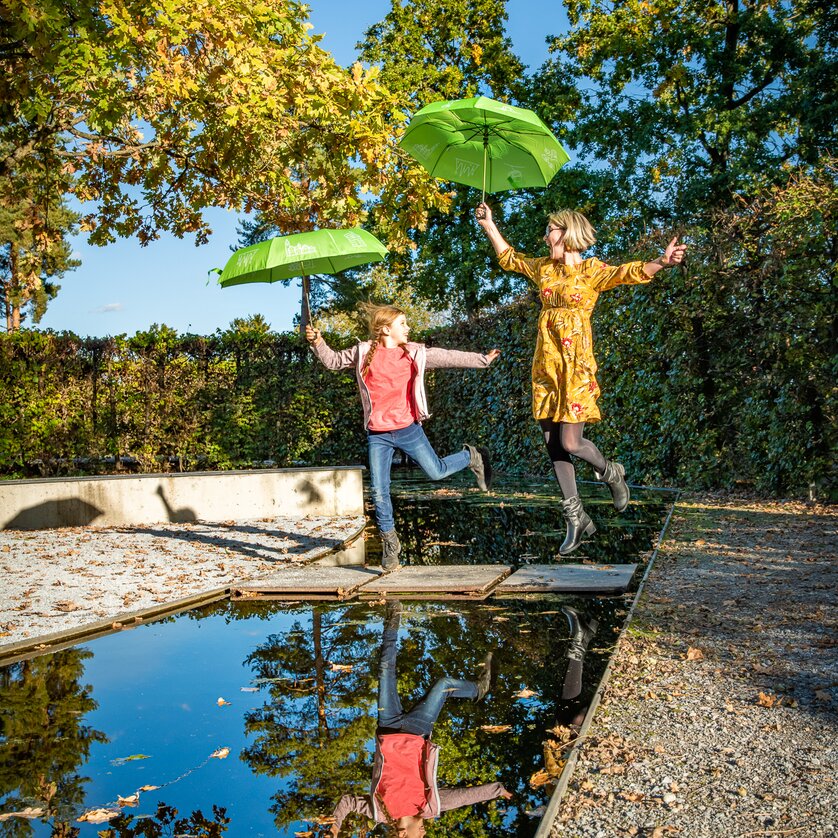 Austrian Sculpture Park | © Region Graz - Mias Photoart A girl and a woman jump over a water surface with green umbrellas. | © Region Graz - Mias Photoart